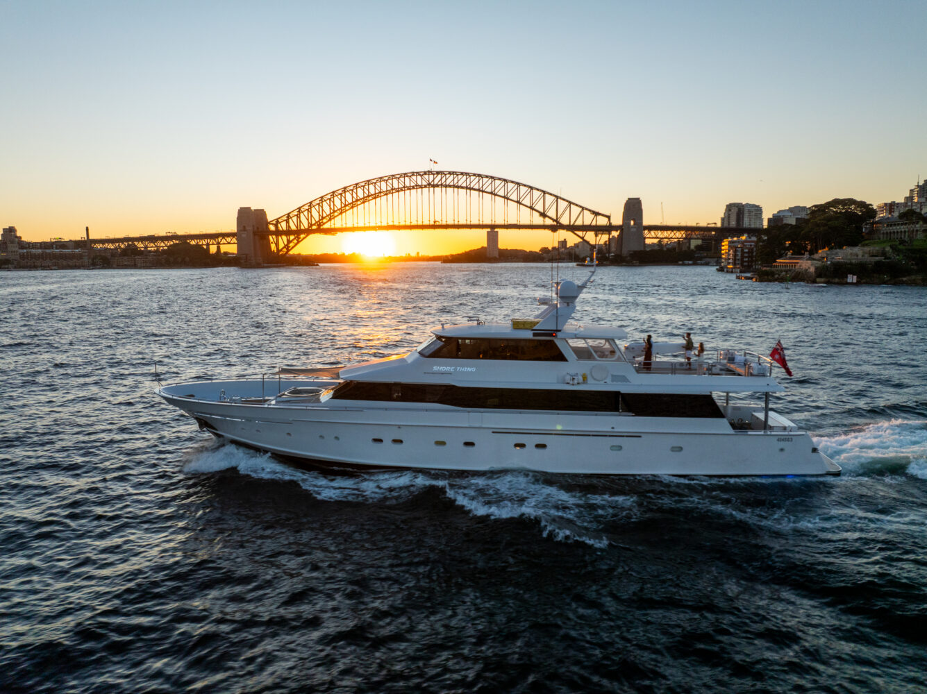 A white yacht cruises on the water at sunset, with the Sydney Harbour Bridge in the background and city buildings visible under a clear sky.