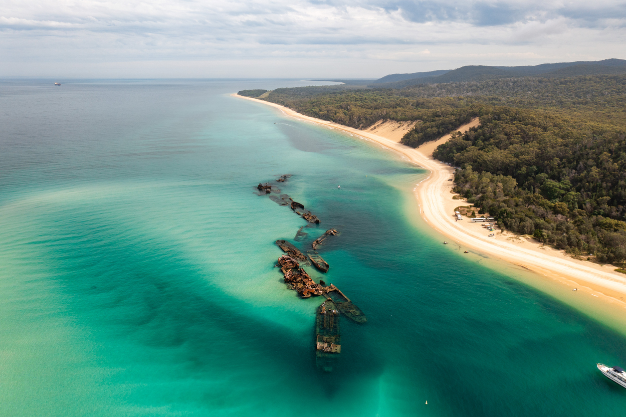 Aerial view of a turquoise sea with visible shipwrecks near a sandy beach bordered by dense green forest under a partly cloudy sky.