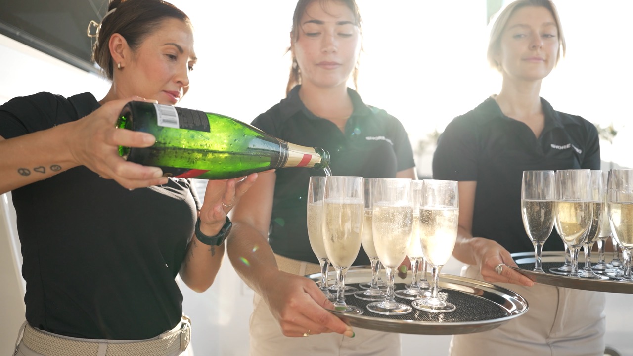 Three women in uniform serve champagne, one pouring into glasses while two hold trays of filled flutes. The sunlight creates a bright, celebratory atmosphere.