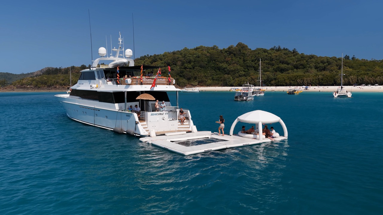 A large white yacht is anchored near a beach with lush green hills. People relax on a floating platform attached to the yacht, some sitting under a shaded canopy. Several other boats are visible near the shore.