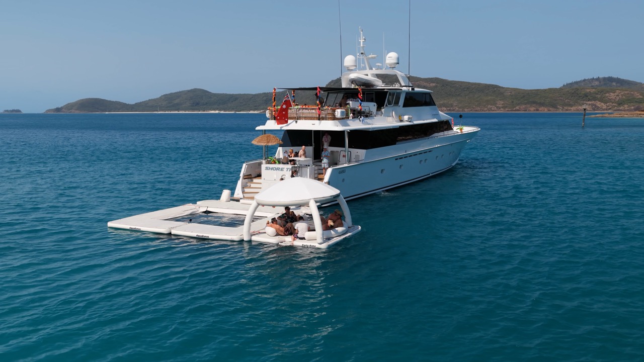 A white yacht is anchored in clear blue water near small green islands. Several people relax on a floating platform attached to the yacht, enjoying the sunny weather.