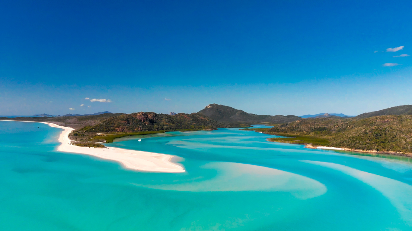 Aerial view of a tropical beach with turquoise water, white sandbanks, and lush green hills under a clear blue sky.