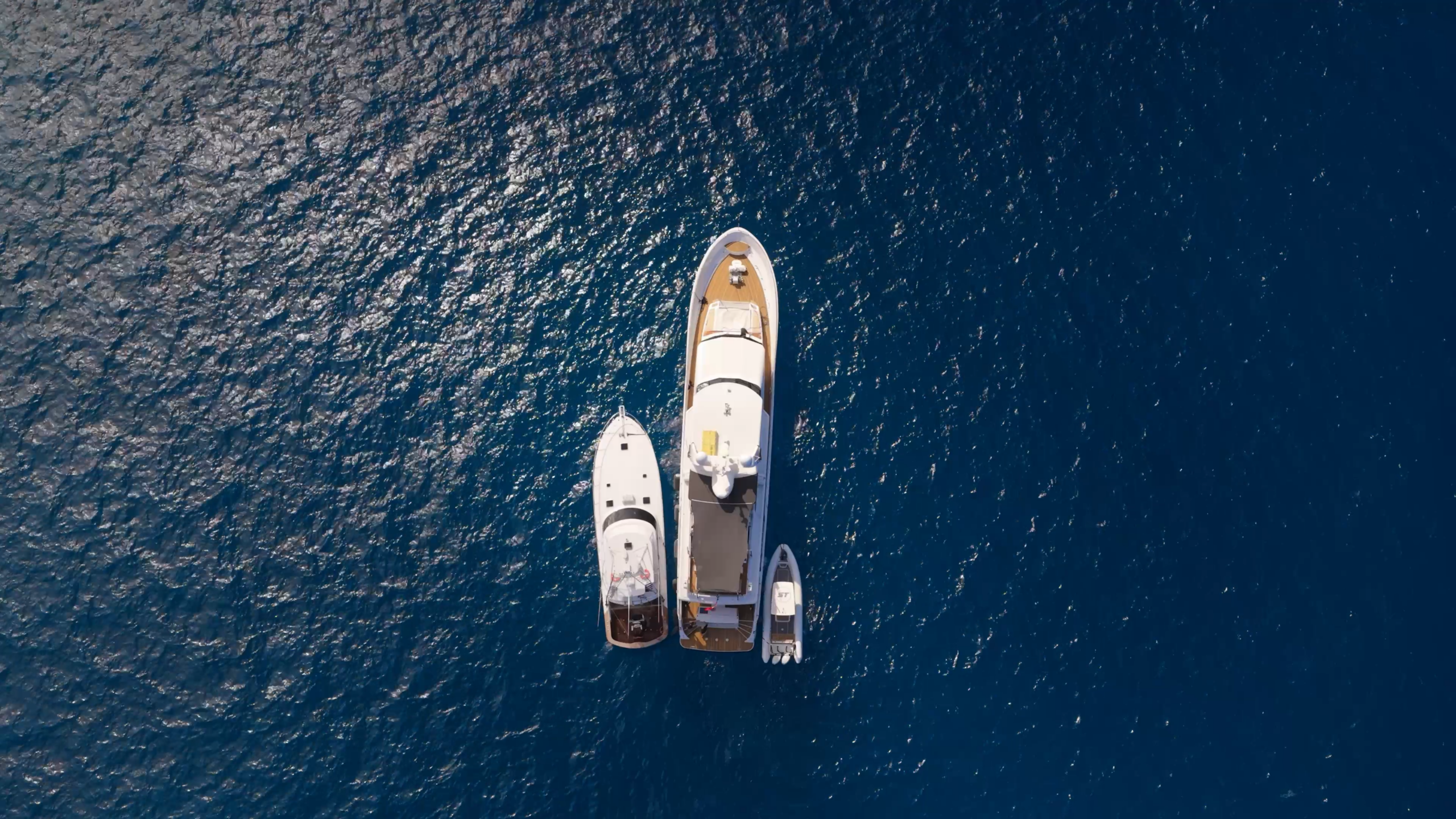Aerial view of three boats, including a large yacht and two smaller vessels, floating side by side on deep blue, sunlit water.