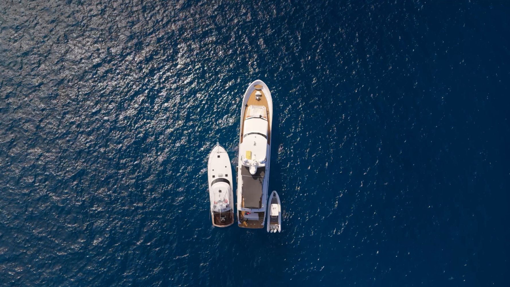 Aerial view of three boats—a large yacht, a medium boat, and a small boat—anchored together in deep blue water with sunlight reflecting off the surface.