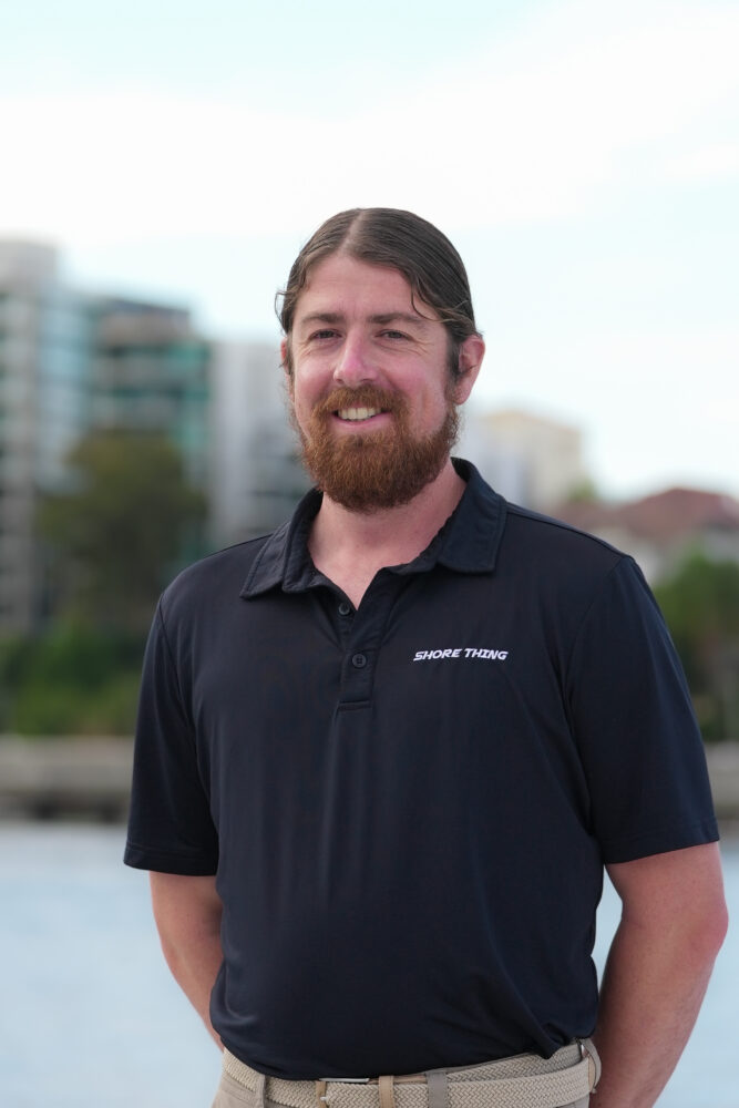 A man with long brown hair and a beard, wearing a black polo shirt that says “SNORK TRKING,” stands outdoors with blurred buildings and trees in the background.