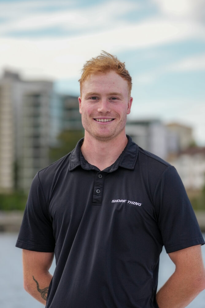 A smiling man with short red hair stands outdoors wearing a black polo shirt that says “SNORE THING.” Buildings and trees appear blurred in the background under a partly cloudy sky.
