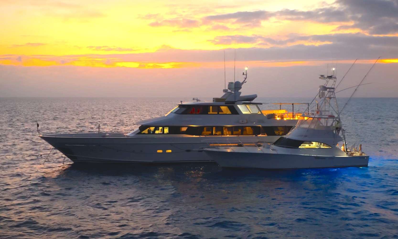 A large, modern yacht with glowing lights is anchored on calm ocean waters at sunset, with orange and yellow hues in the sky and gentle waves surrounding the boat.