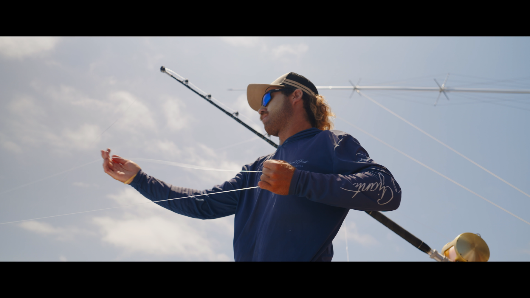 A person in a blue shirt, hat, and sunglasses is fishing, holding fishing line with a pole behind them under a bright, partly cloudy sky.