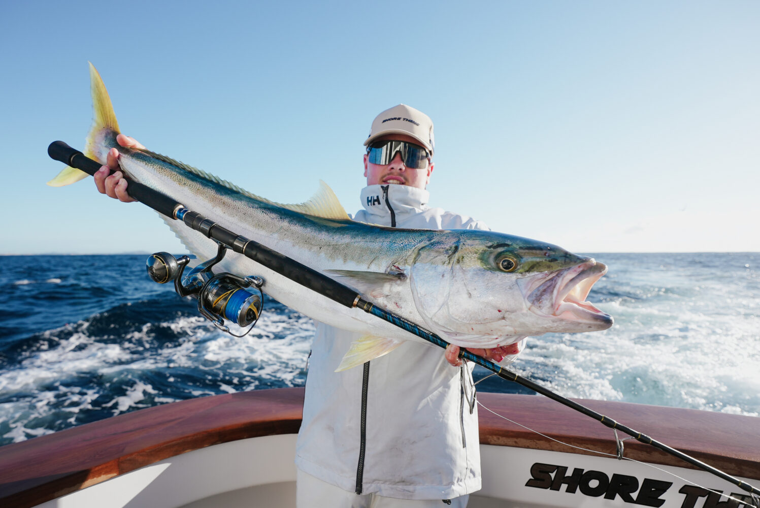 Person in white jacket and cap holding a large fish with a fishing rod on a boat, ocean waves and clear sky in the background.