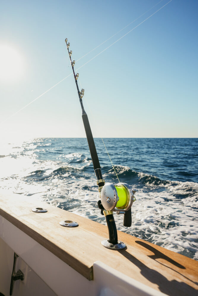 A fishing rod with a bright green reel is mounted on a boat, facing the ocean. Waves splash behind the boat under a clear blue sky, with sunlight shining from the left.