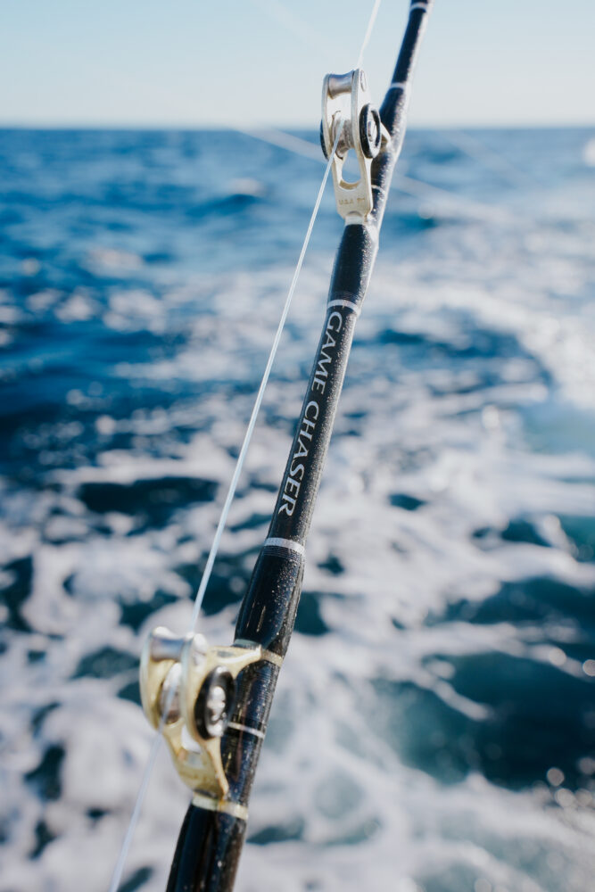 A close-up of a fishing rod labeled “Game Chaser” on a boat, with ocean waves and blue sky visible in the background.