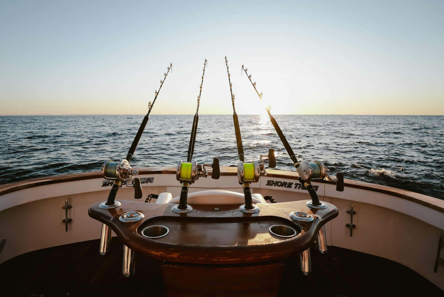 Three fishing rods are set up at the back of a boat on the open ocean at sunset, with calm water and a clear sky in the background.