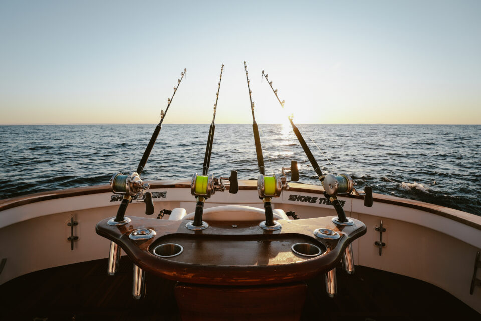 Three fishing rods are set up at the back of a boat on the open ocean at sunset, with calm water and a clear sky in the background.