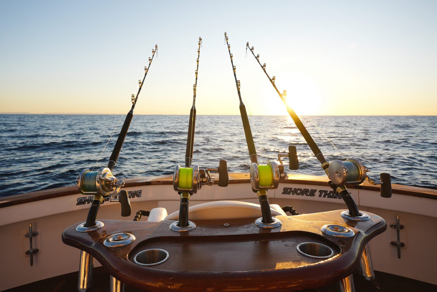Three fishing rods are mounted on the back of a boat at sunrise or sunset, with the ocean and a bright sun visible on the horizon.