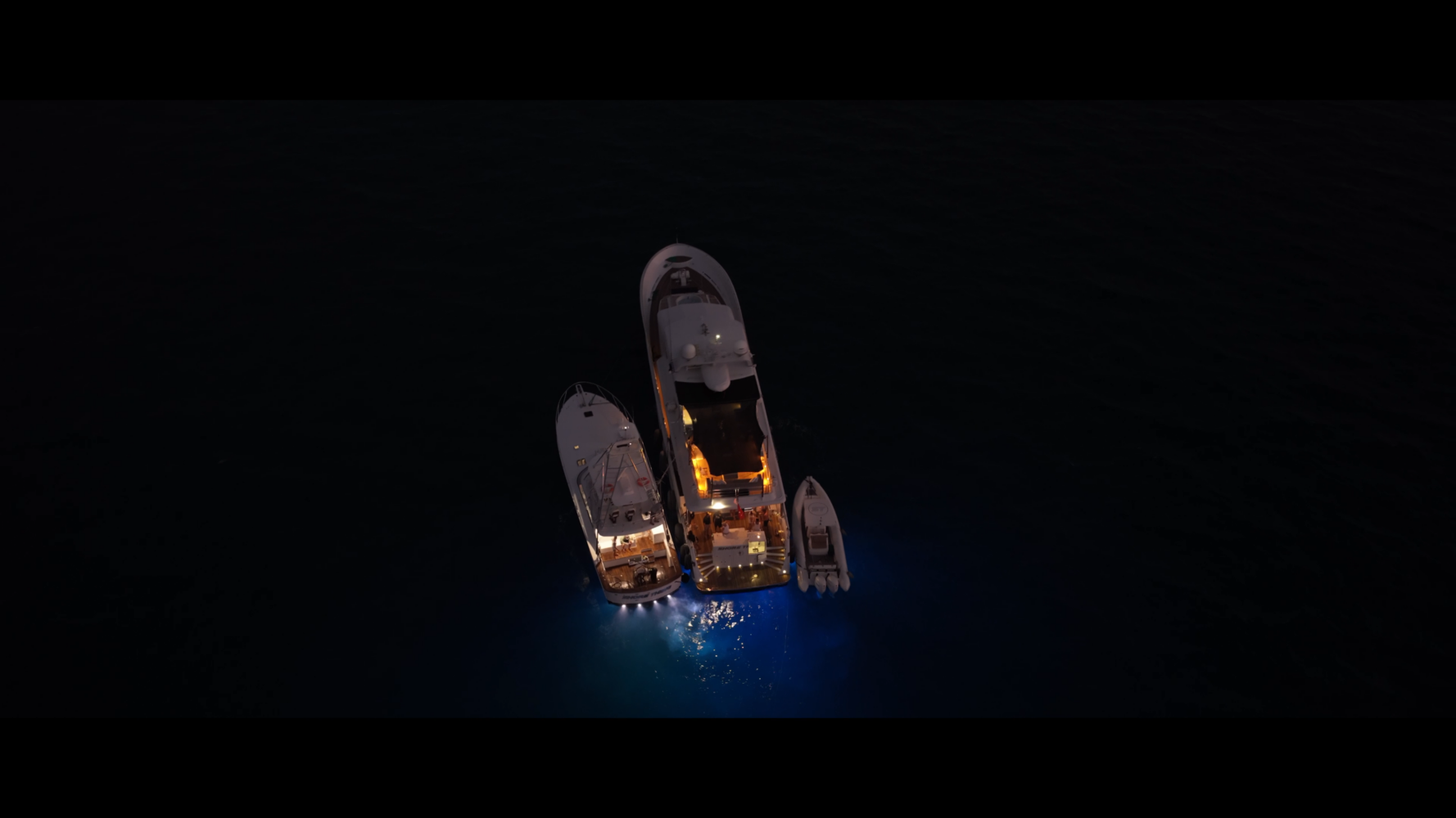Aerial view of two yachts and a small boat anchored side by side at night, illuminated by warm lights reflecting on the dark water.