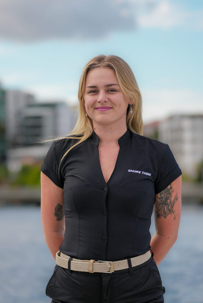 A woman with blonde hair, wearing a black “Shore Thing” shirt and beige belt, stands outdoors by the water with buildings and a cloudy sky in the background. She is smiling with her hands behind her back.