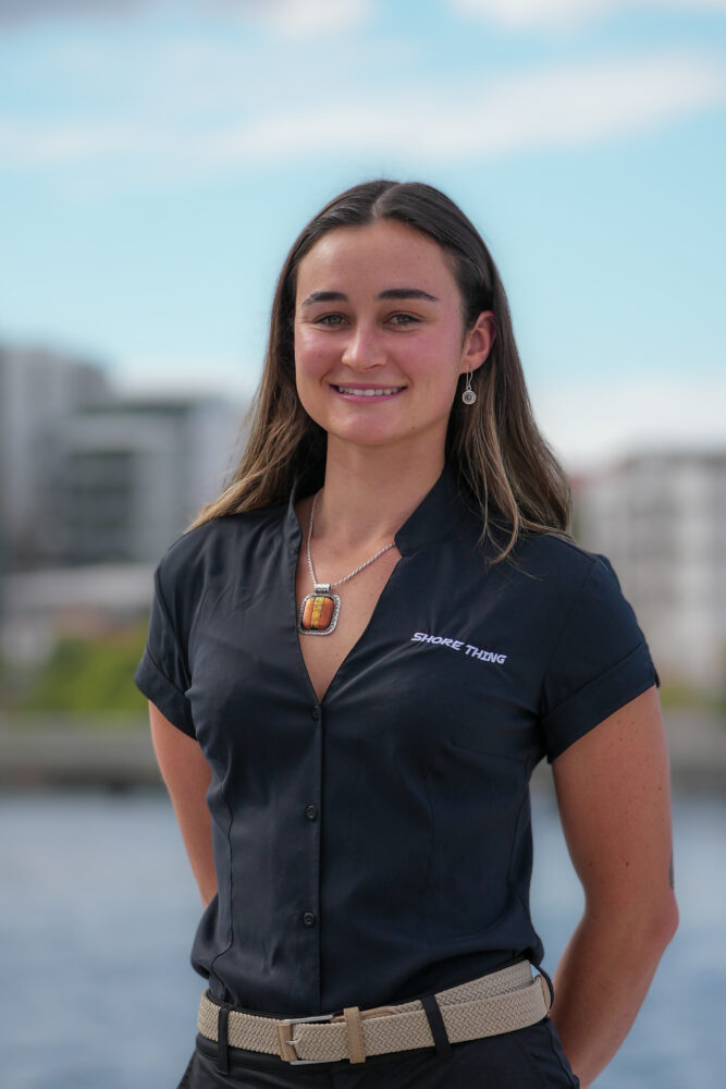 A woman with long brown hair, wearing a black short-sleeved shirt and a pendant necklace, stands outdoors with a blurred cityscape and water in the background. She is smiling at the camera.
