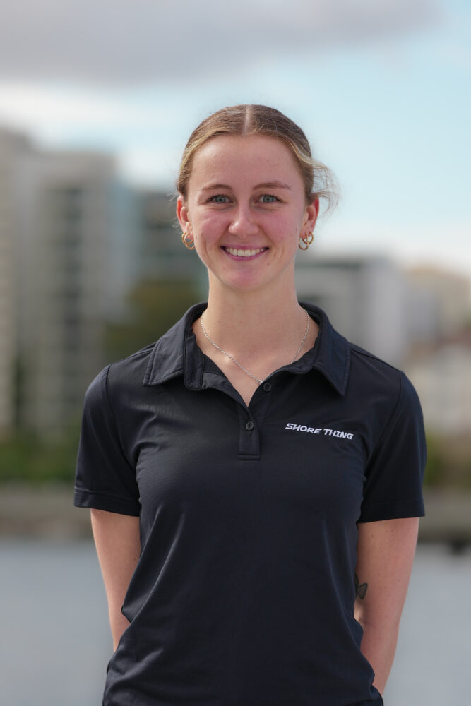 A young woman with light brown hair tied back smiles outdoors, wearing a black collared shirt with “SHORE THING” written on it. The background is blurred with buildings and a partly cloudy sky.