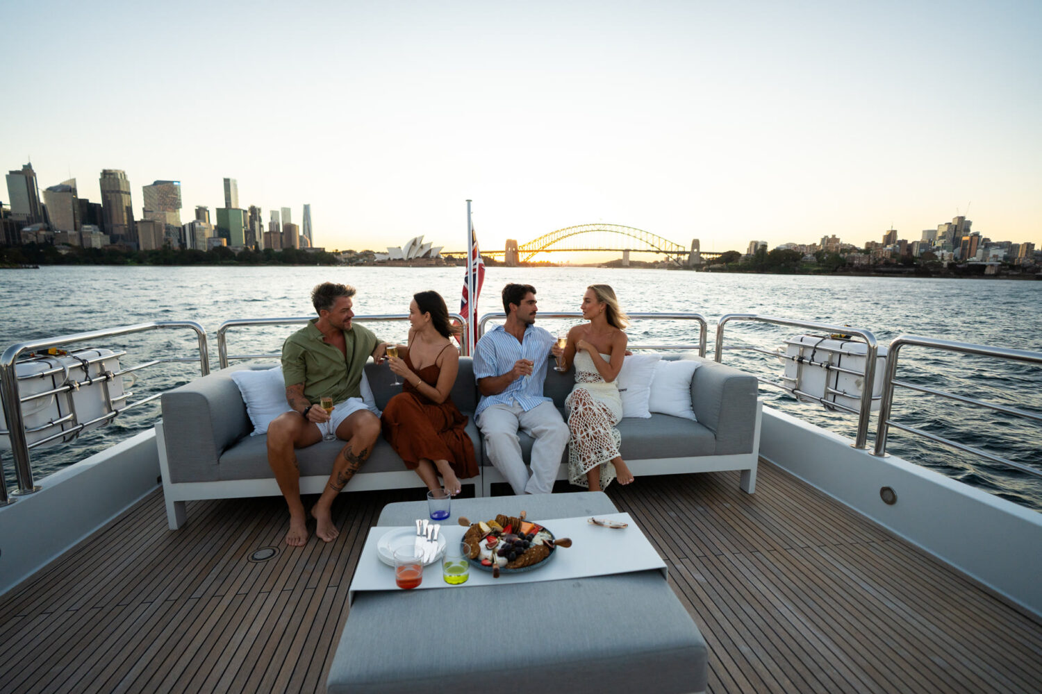 Four people sit on a yacht’s sofa, talking and holding drinks. A platter of food is on the table in front of them. The Sydney Opera House and Harbour Bridge are visible in the background at sunset.