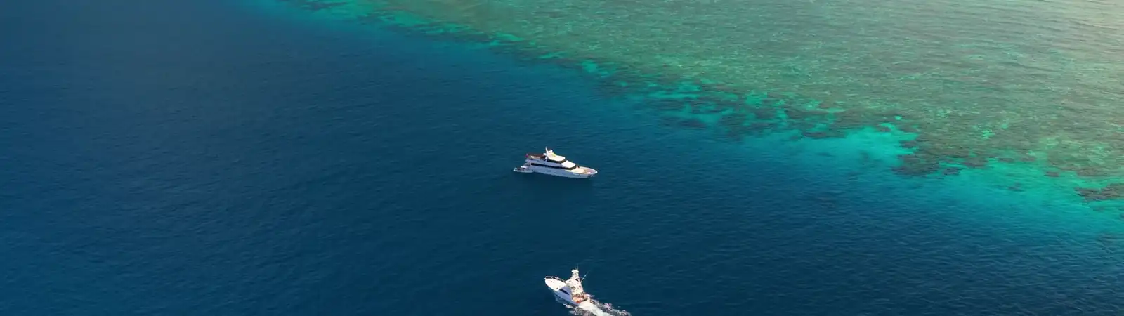 Two boats float on deep blue water near a colorful coral reef. The reef’s turquoise and green hues contrast with the darker open sea, showing the boundary between shallow and deep water from an aerial perspective.