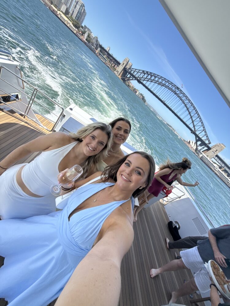 Three women in light-colored dresses smile for a selfie on a boat with Sydney Harbour Bridge and city buildings in the background. Other people are on the deck enjoying the sunny day by the water.