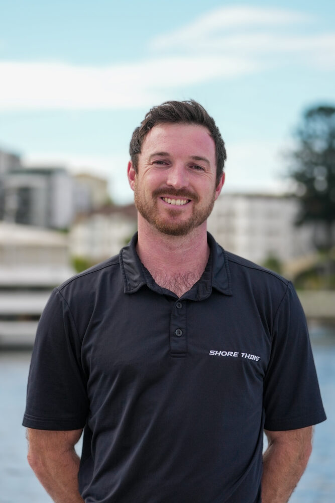 A man with short brown hair and a beard smiles, wearing a black polo shirt that says “Shore Thing.” He stands outdoors with blurred buildings and water in the background under a partly cloudy sky.