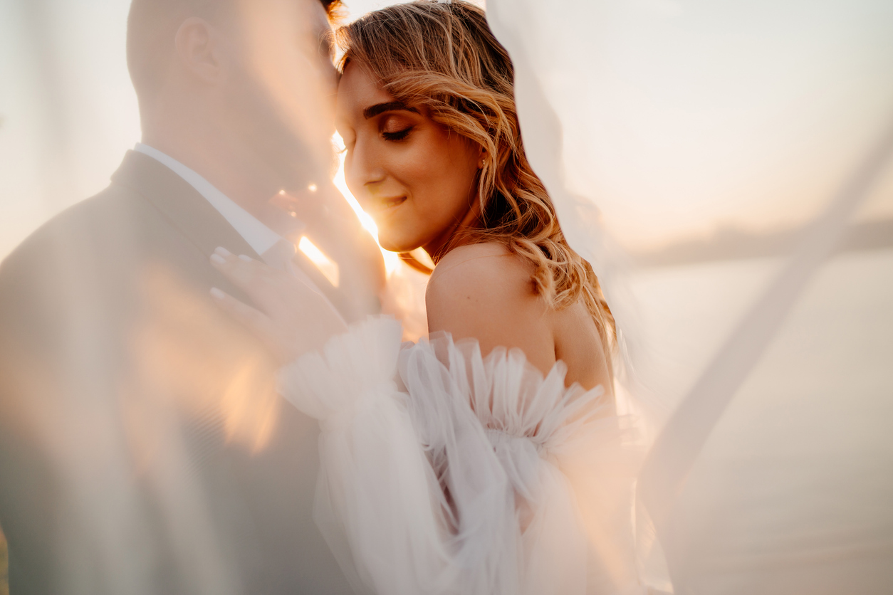 A woman in a sheer, off-shoulder dress embraces a man in a suit at sunset, both softly lit with white fabric flowing around them, creating a dreamy, romantic atmosphere.