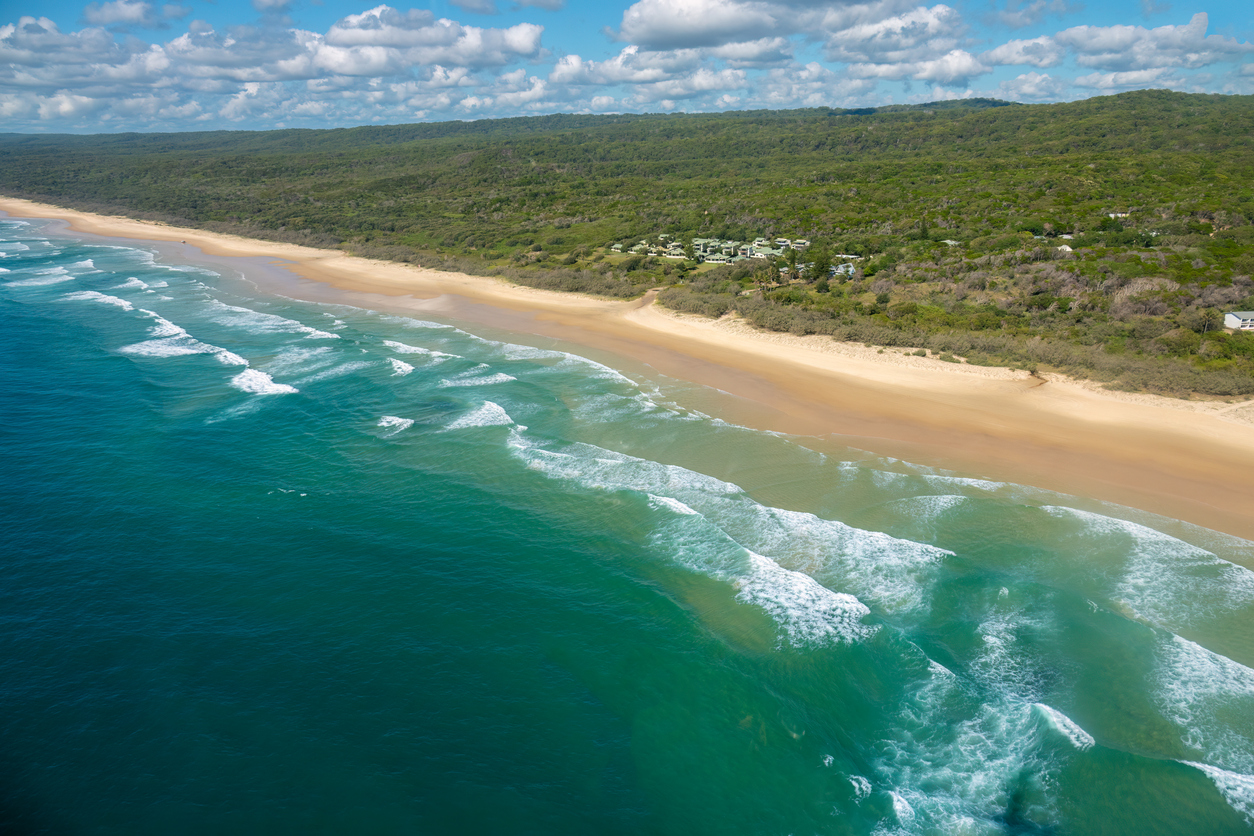 Aerial view of a sandy beach with turquoise waves, bordered by dense green forest under a partly cloudy sky.