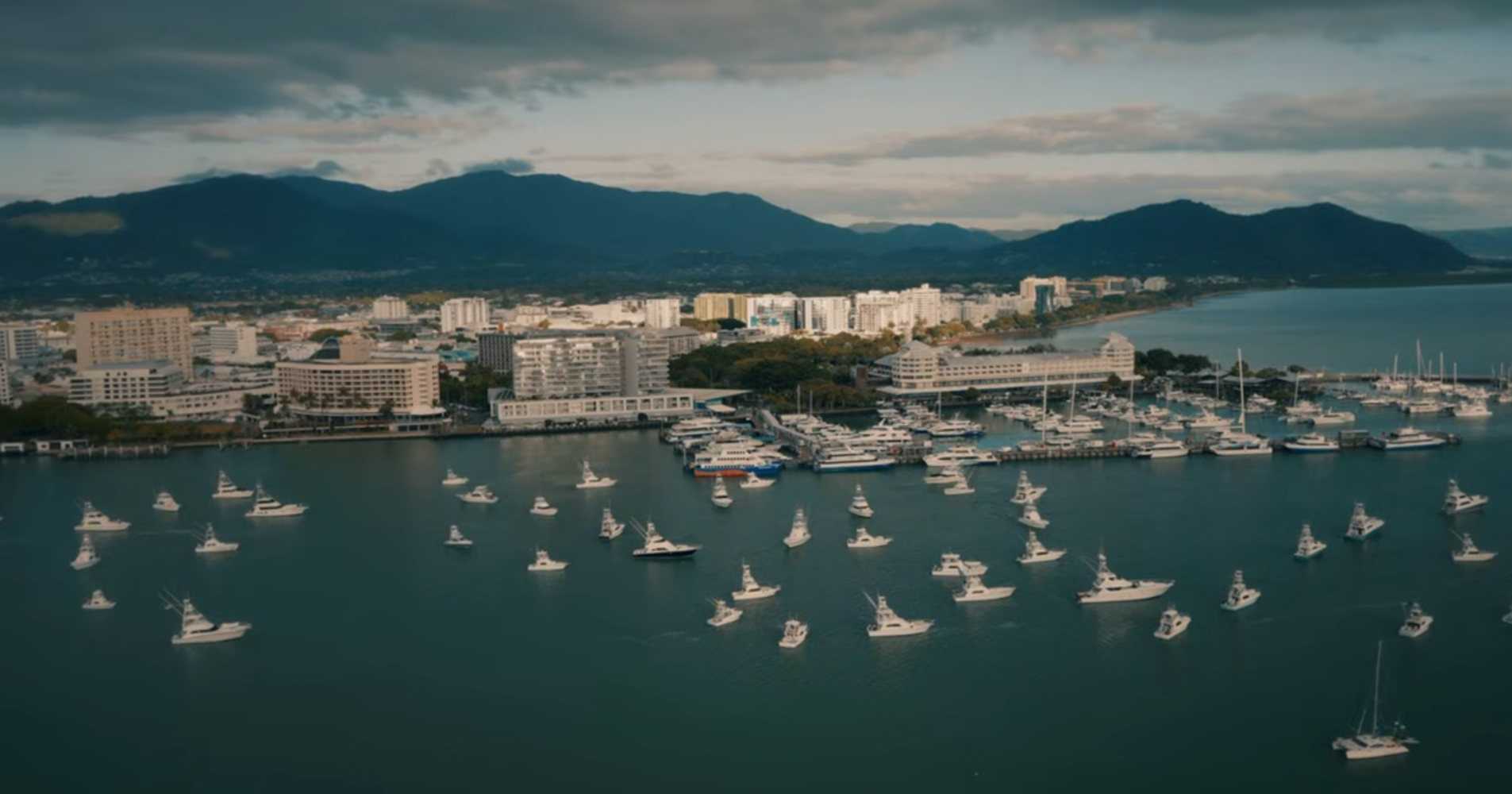 Aerial view of a marina filled with boats in front of a coastal city with mountains in the background.