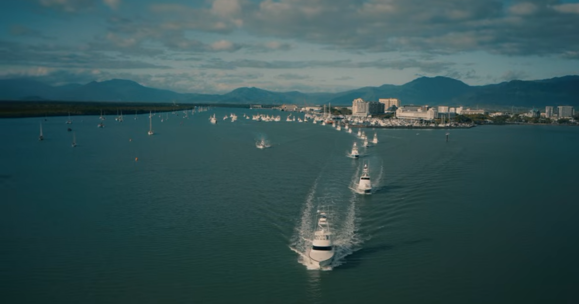 A line of motor yachts travels across a bay toward a coastal city with mountains in the background.
