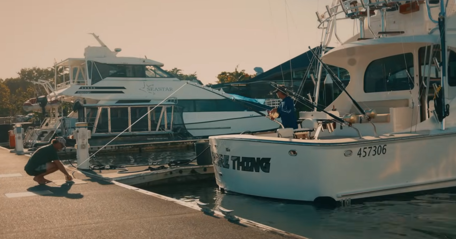 Two people at a marina, one kneeling on the dock securing a rope and the other standing on a boat named “Sure Thing.”.
