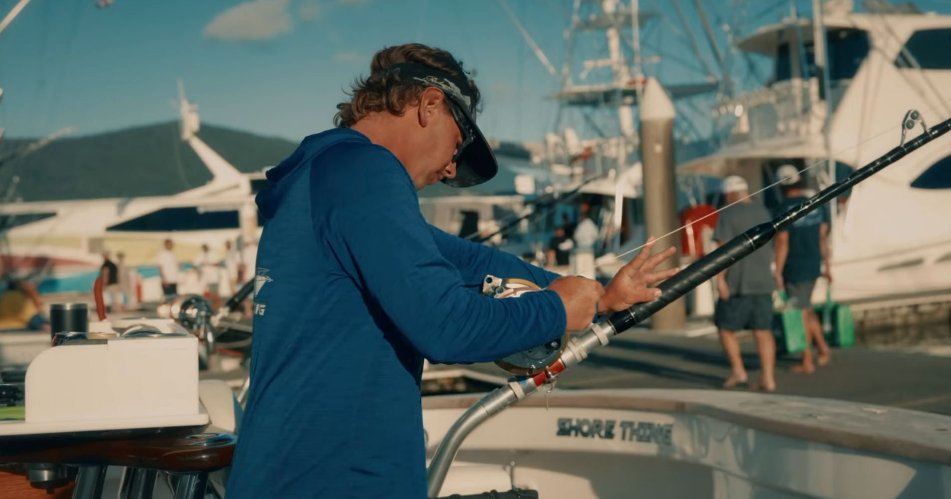A man in a blue long-sleeved shirt reels in a fishing line on a boat at a marina.
