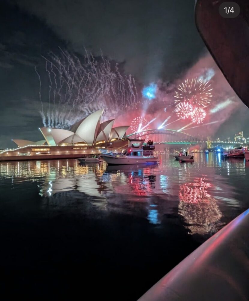 Fireworks explode above the Sydney Opera House and Harbour Bridge, with game fishing boats on the water reflecting the lights.