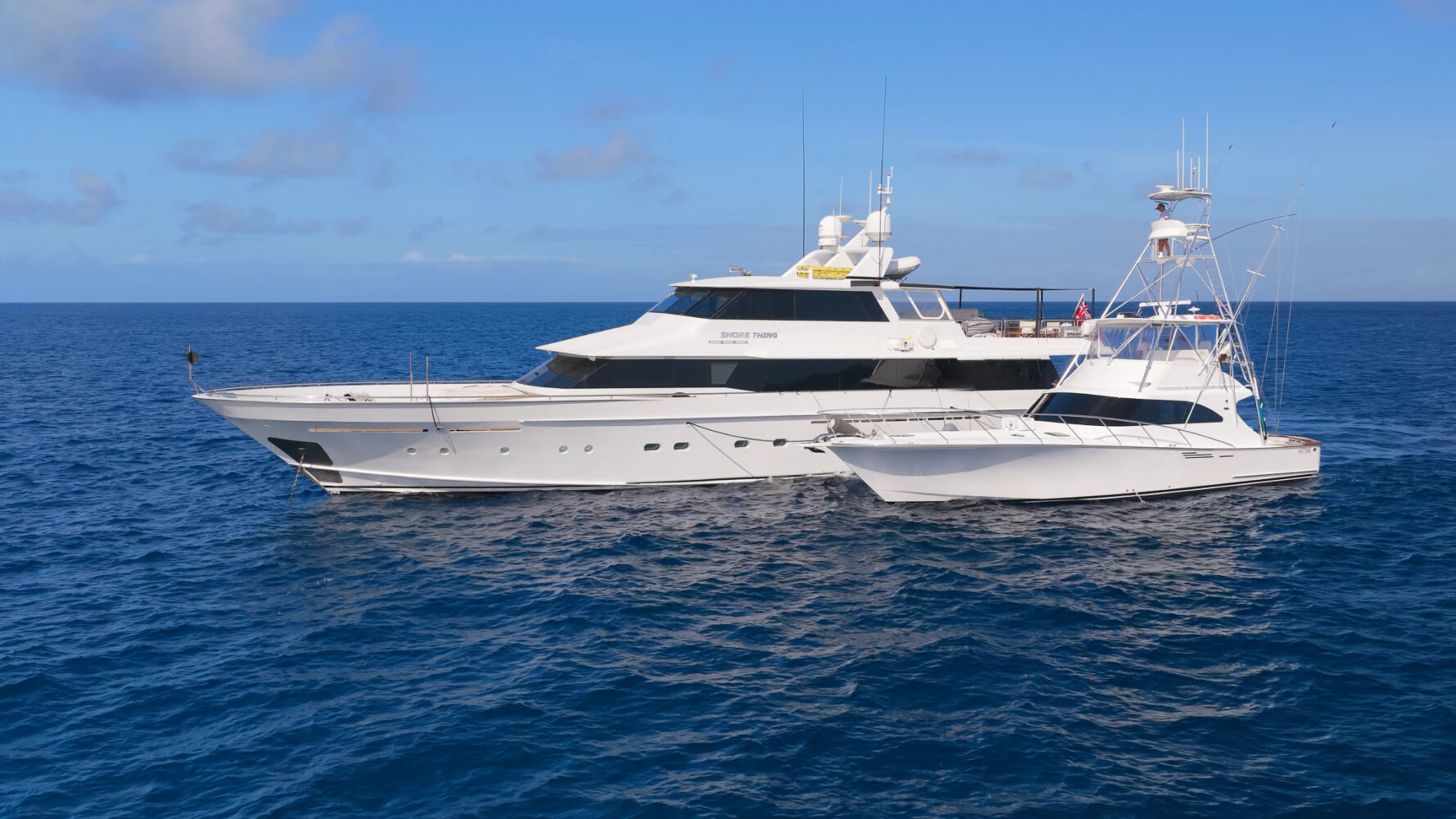 A large white motor yacht named “Shore Thing” docked next to a smaller white fishing boat in the open ocean.