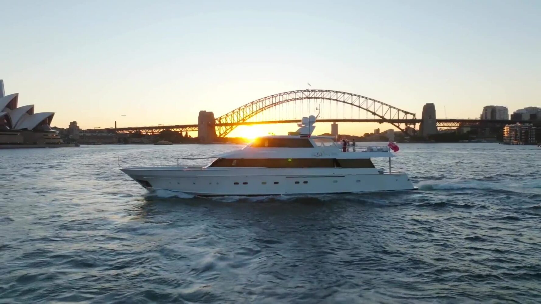 A white yacht sails on the water in front of the Sydney Harbour Bridge and Opera House at sunset.