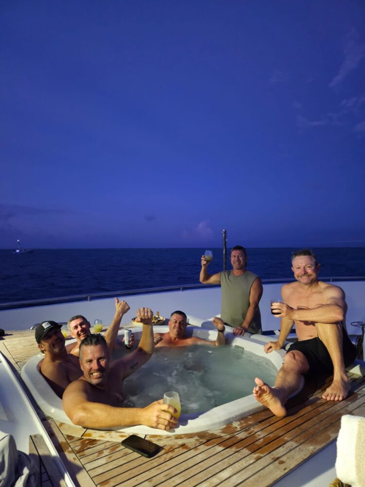 Five men relaxing in a hot tub on a boat during a corporate Christmas party in Sydney, holding drinks and smiling at the camera at twilight.