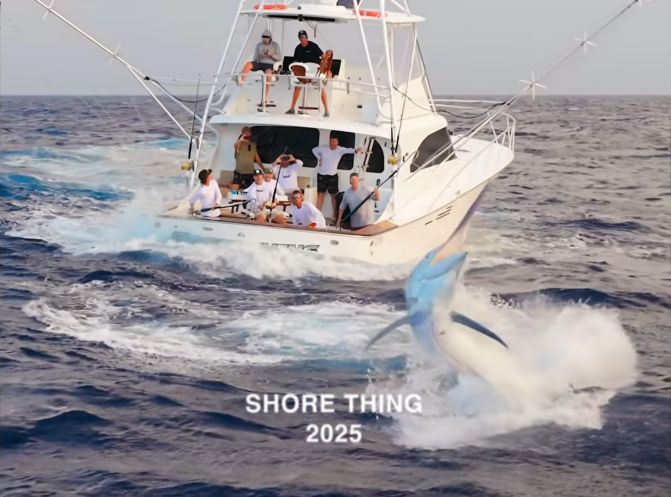 A marlin leaps from the ocean behind a fishing boat occupied by a group of people, with the text “SHORE THING 2025” overlaid at the bottom.